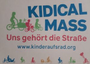 Das Bild zeigt ein Plakat mit dem Titel "KIDICAL MASS" in großen blauen Buchstaben. Darunter steht in roten Buchstaben "Uns gehört die Straße". Die Website "www.kinderaufsrad.org" ist in blauer Schrift angegeben. Das Plakat zeigt Silhouetten von Kindern und Erwachsenen, die auf Fahrrädern fahren. Es gibt verschiedene Farben, darunter Grün, Rot, Blau und Orange, die die Silhouetten darstellen. Die Silhouetten zeigen Familien, die zusammen auf Fahrrädern fahren, einschließlich eines Kindertransporters. Am unteren Rand des Plakats sind weitere Silhouetten von Kindern und Erwachsenen, die auf Fahrrädern fahren, zu sehen. Einige der Silhouetten haben Ballons.
