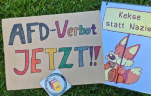 Zwei Protestschilder mit der Aufschrift "AfD Verbot jetzt!" und "Kekse statt Nazis" (Fuchs-Motiv von Fuchskind) liegen auf einem grünen Rasen. Ein kleinerer Aufkleber mit der Abkürzung „GG“ (Grundgesetz) und dem Hinweis auf Artikel 1 ist ebenfalls auf dem Schild angebracht.