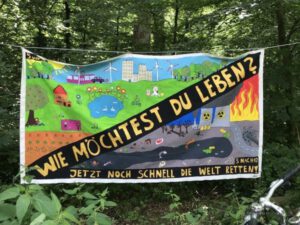 Ein handgemaltes Banner hängt zwischen Bäumen. Es zeigt zwei gegensätzliche Szenarien: Eine Hälfte zeigt eine idyllische Landschaft mit Häusern, Windrädern, einem See und glücklichen Menschen. Die andere Hälfte zeigt eine dystopische, von Umweltverschmutzung und Feuer zerstörte Landschaft. Der Text auf dem Banner lautet „Wie möchtest du leben?“, mit dem Zusatz „Jetzt noch schnell die Welt retten“. Das Banner ist bunt und kindlich gezeichnet.