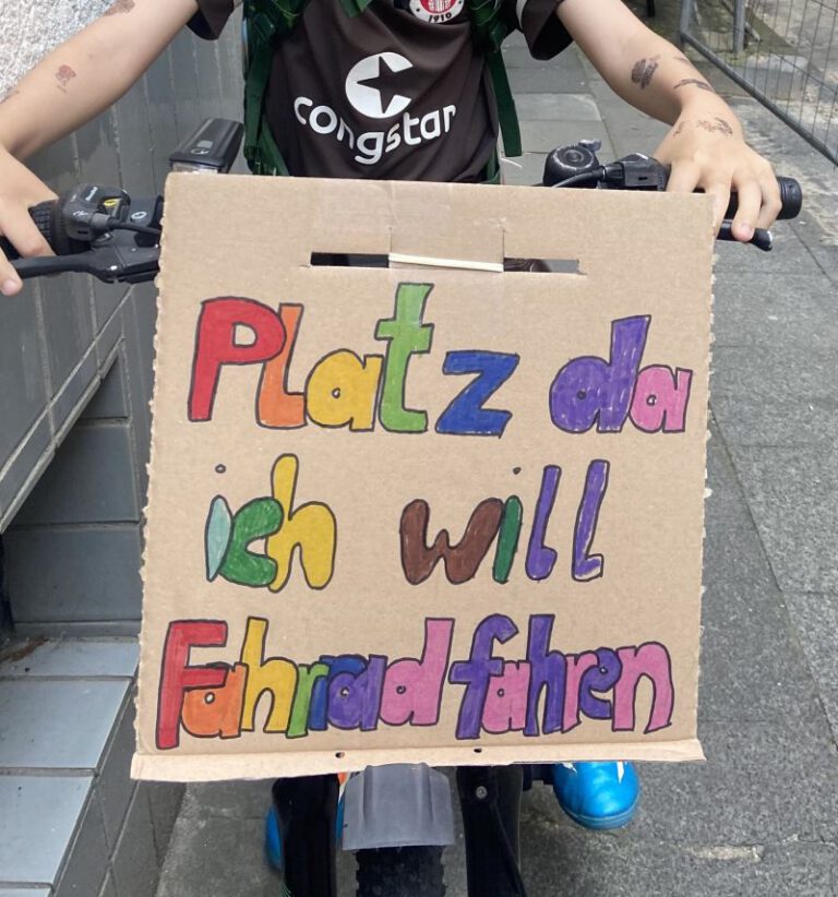 Ein Mensch auf einem Fahrrad mit dem vorne montierten Schild: Platz da! Ich will Fahrrad fahren