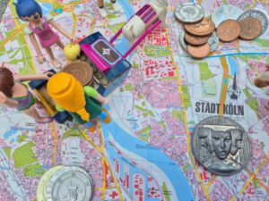 Ein alter Stadtplan von Köln (auf Papier!) Darauf stehen Playmobildfiguren, die einen von einem Pferd gezogenen Schlitten umringen. Auf dem SChlitten liegen alter Taler. Auf dem Stadtplan sind ebenfalls einige Münzen verteilt.
