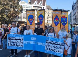 Das Foto zeigt eine Demonstration in Köln. Mehrere Teilnehmer halten große Schilder mit Botschaften und Bildern. Im Vordergrund ist das Team der Verkehrswende Köln mit der Forderung ObenBleiben - kein Ost-West-Tunnel zu sehen. Im Hintergrund sind Schilder mit stilisierten afrikanische Masken . Auf den Schildern steht "Alarm", "Auf", "Planet Erde".