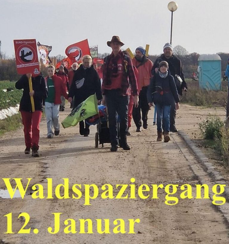 Das Bild zeigt eine Gruppe von Menschen, die auf einem Feldweg spazieren geht. Bei diesem Protestmarsch sind Schilder zu sehen. Diese zeigen Symbole und Schriftzüge aus dem Braunkohlewiderstand. Unten im Bild steht in gelben Lettern „Waldspaziergang 12. Januar“.