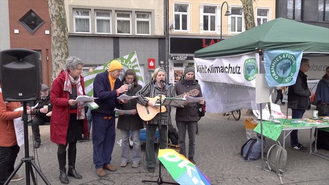 "Wählt Klimaschutz" - Infostand und Kundgebung am 22.02.25 auf dem Chlodwigplatz in Köln Im Bild: Teile des Kölner Klimachors singen mit Gitarrenbegleitung. Auf der rechten Bildseite steht der Infopavillion mit grünem Dach. Im Bild der Infopavillion mit grünem Dach. An den Seiten ist das Banner "Wählt Klimaschutz" sowie die Fahnen von Parents und GRannies for Future Köln befestigt. Foto: Ulla Kellerwessel