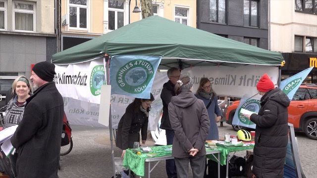 "Wählt Klimaschutz" - Infostand und Kundgebung am 22.02.25 auf dem Chlodwigplatz in Köln Im Bild der Infopavillion mit grünem Dach. An den Seiten ist das Banner "Wählt Klimaschutz" sowie die Fahnen von Parents und GRannies for Future Köln befestigt. Vorne sind zwei Tische mit Informationsmaterialien aufgebaut. Mehrere Menschen fürhen intensive Gespräche. Foto: Ulla Kellerwessel