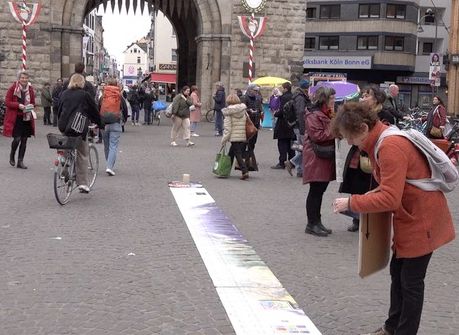 "Wählt Klimaschutz" - Infostand und Kundgebung am 22.02.25 auf dem Chlodwigplatz in Köln - Im Bild: vor der gut belebten Straße liegt der 25 Meter lange Klimazeitstrahl. Eine Passantin betrachtet interessiert Details Foto: Ulla Kellerwessel