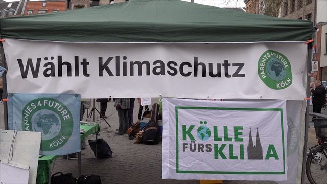 "Wählt Klimaschutz" - Infostand und Kundgebung am 22.02.25 auf dem Chlodwigplatz in Köln Im Bild eine Seitenansicht des Infopavillions mit grünem Dach. Das Banner "Wählt Klimaschutz" sowie die Fahnen von Parents und GRannies for Future Köln sind daran befestigt. Ferner die "Kölle fürs Klima" Fahne. Foto: Ulla Kellerwessel