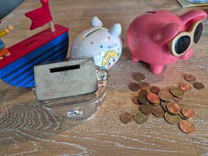 Auf einem Holztisch stehen vier verschiedene Spardosen. Links im Bild ist ein blauer, als Schiff gestaltete Halbscheibe aus Holz. Das Oberdeck ist rot lackiert, dort in Mittig der Geldschlitz. Links steht eine Holzfigur, rechts ist eine rote Fahne. In der Mitte hinten ist ein klassisches weißes Porzellanschwein. Es ist mit Punkten und einem blonden Kind bemalt. Auf der rechten Seite ist ein rosa farbenes Porzellanschwein mit Sonnenbrille. Im Vordergrund ist ein aus Metall gefertigtes SParhaus. Rund um den Einwurfschlitz sind deutliche Kratzspuren zu sehen. Rechts vorne liegt ein Haufen Kleingeld.