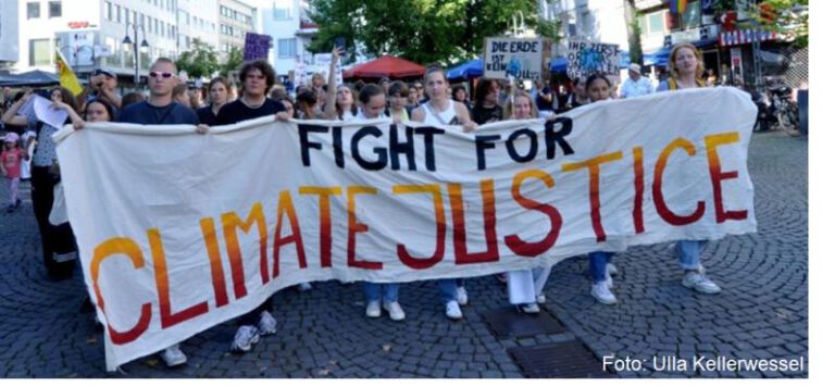 Das Bild zeigt eine Gruppe von Menschen, die am Kölner Klimastreik teilnehmen. Sie halten ein großes Banner hoch, auf dem in großen, farbenfrohen Buchstaben "FIGHT FOR CLIMATE JUSTICE" steht. Die Menschen sind in einer städtischen Umgebung zu sehen, mit gepflasterten Straßen und Gebäuden im Hintergrund. Viele der Leute sind jung und tragen leichte, sommerliche Kleidung. Einige halten kleinere Schilder mit weiteren Botschaften, wie "DIE ERDE IST KEIN WARENLAGER" und "IHR ZERSTÖRT WAS UNS ALLEN GEHÖRT". Foto: Ulla Kellerwessel