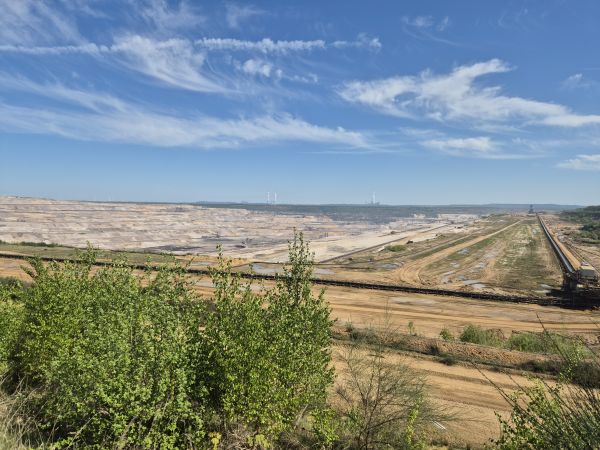Das Bild zeigt den Tagebau Hambach, ein gewaltiger Braunkohletagebau unter einem klaren, blauen Himmel mit einigen weißen Wolken. Im Vordergrund sind grüne Büsche und Sträucher zu sehen, die einen Kontrast zu der trockenen, erodierten Landschaft bilden. In der Mitte des Bildes erstreckt sich eine riesige, offene Grube. Der Tagebau wird von einem langen, breiten Förderband durchquert, das sich von rechts nach links erstreckt. Im Hintergrund sind Windräder und Kohlekraftwerke zu sehen.