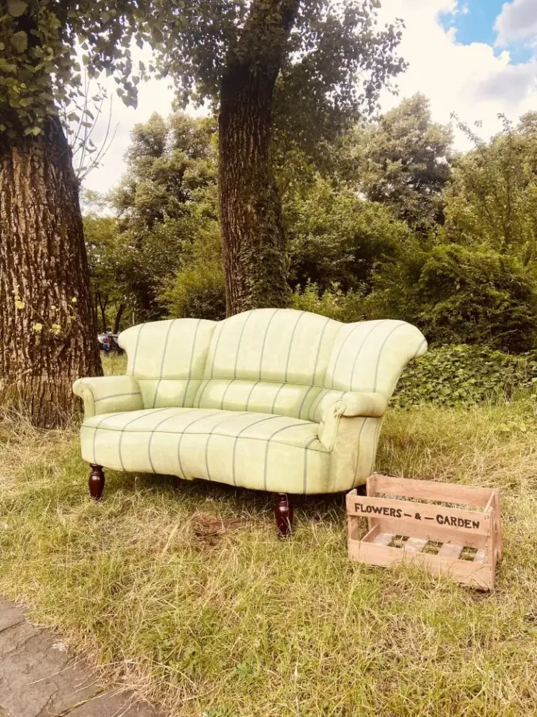 Am Wegesrand steht ein weißliches Sofa auf grünem Gras. Daneben steht eine Holzkiste mit der Aufschrift "Flowers & Garden". Im Hintergrund sind zwei Lindenbäume sowie Sträucher zu sehen. Der Himmel ist teils bewölkt, rechts oben ist ein wenig blauer Himmel sichtbar.