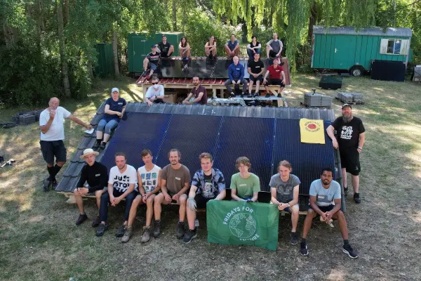 Das Bild zeigt eine Gruppe von jungen Menschen und Erwachsenen vor zwei kleinen Testdächern mit Solarpanelen, einige halten ein grünes Banner mit der Aufschrift 'Fridays for Future' in den Händen. Die Dächer stehen auf einer kurz gemähten Wiese. Im Hintergrund sind grüne Bauwagen, Paletten mit Material und grüne Bäume zu sehen.