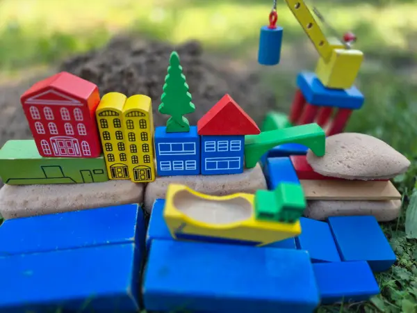 Eine bunte Landschaft aus Holzspielzeug: Häuser stehen auf Schwämmen, im Vordergrund ist ein Fluss mit blauen Holzklötzen gebaut. Rechts hinten ist ein Kran aus Holz mit einer Last. Auf dem Fluss befindet sich ein gelbes Boot.